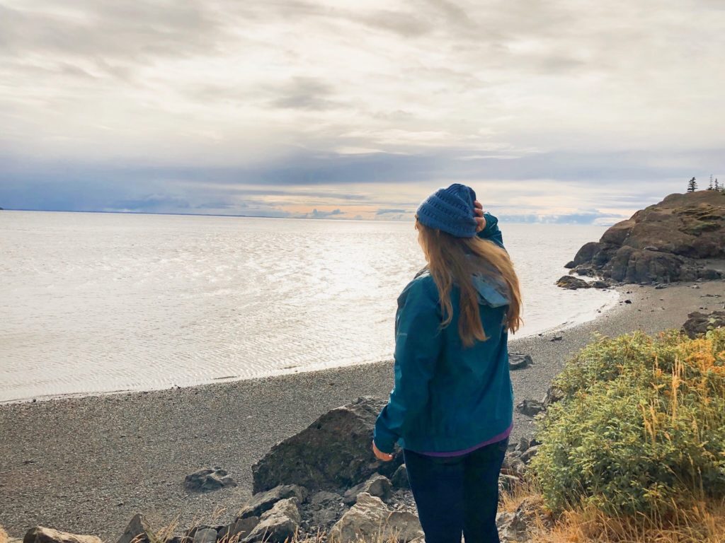 girl in blue jacket and beanie standing on shoreline of turnigan arm waterway looking at ocean and sunset