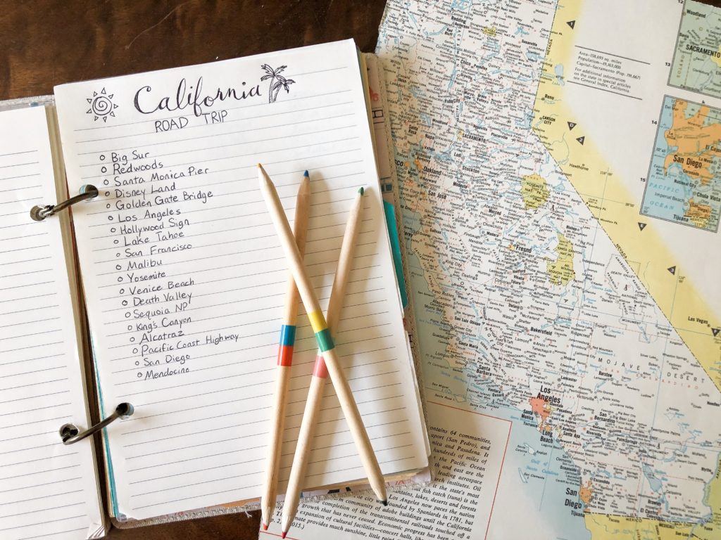 california road trip bucket list in travel journal with wooden colored pencils and antique map of california on wood surface