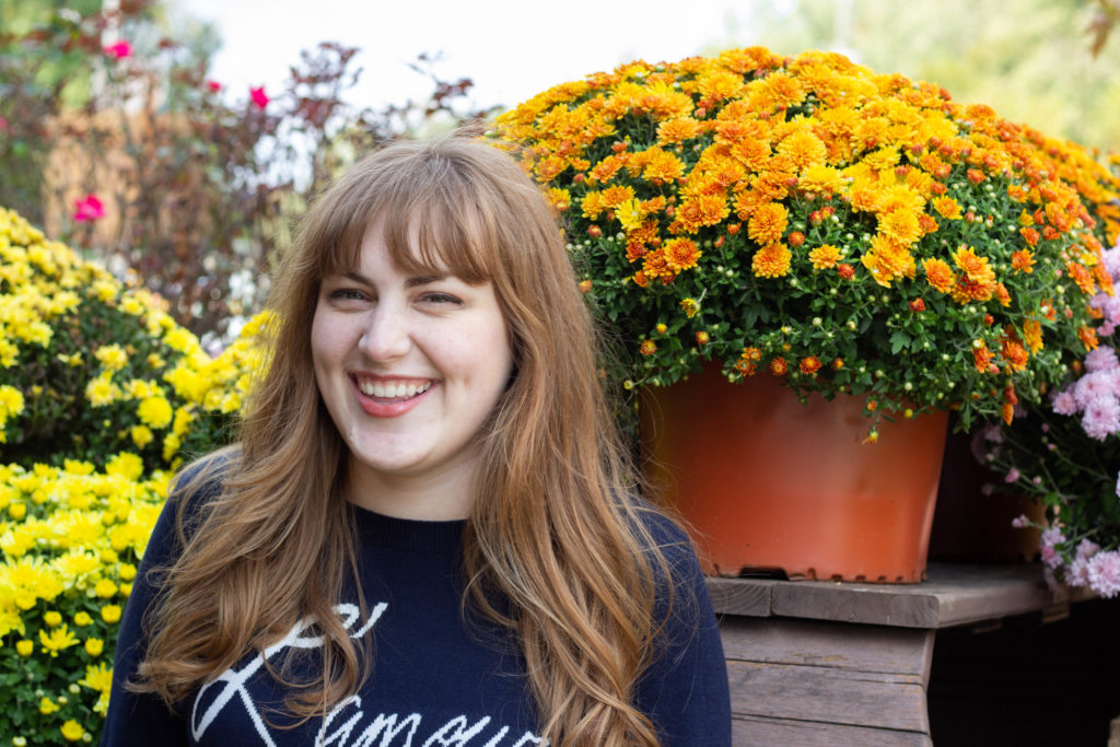 girl laughing at fall photoshoot in navy sweater with white words, orange and yellow mums in background