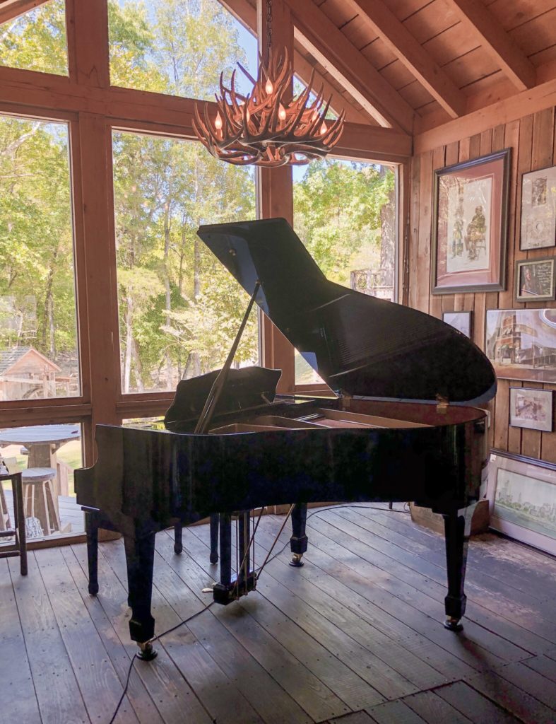 black grand piano in front of big windows and chandelier with rustic decor in the Outpost in pickwick savannah tennessee